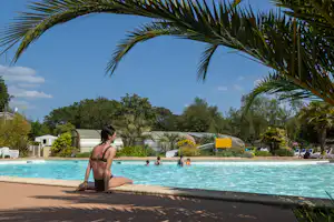 Frau am Camping-Pool im Vordergrund, Palmen und Liegen am Beckenrand, im Hintergrund Zelte und Bäume.