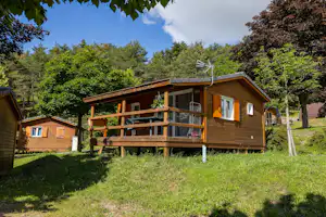 Holzbungalow mit Veranda und Geländer, daneben ein kleiner Schuppen, umgeben von Bäumen und Rasenfläche.