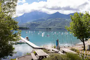 Holzsteg am See mit Booten, Blick auf die Berge und Tische am Campingplatz zwischen Bäumen.