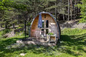 Geodätisches Zelt mit Holzterrasse im Wald, davor Stühle und Tisch, seitlich ein Sonnenschirm.