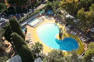 Blick auf den Camping-Pool mit Liegen, Sonnenschirmen und Palmen im Garten, umgeben von Hecken und Wegen.