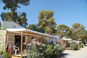 Mobilheime mit Terrasse und Markise, umgeben von blühenden Sträuchern und hohen Bäumen vor blauem Himmel.