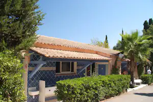 Campinghaus mit Ziegeldach, Fliesenwänden und kleinem Fenster, umgeben von Büschen und Palmen im Garten.