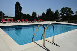 Camping-Pool mit Treppe aus Edelstahl und roten Liegen am Beckenrand, im Hintergrund Bäume und Gebäude.