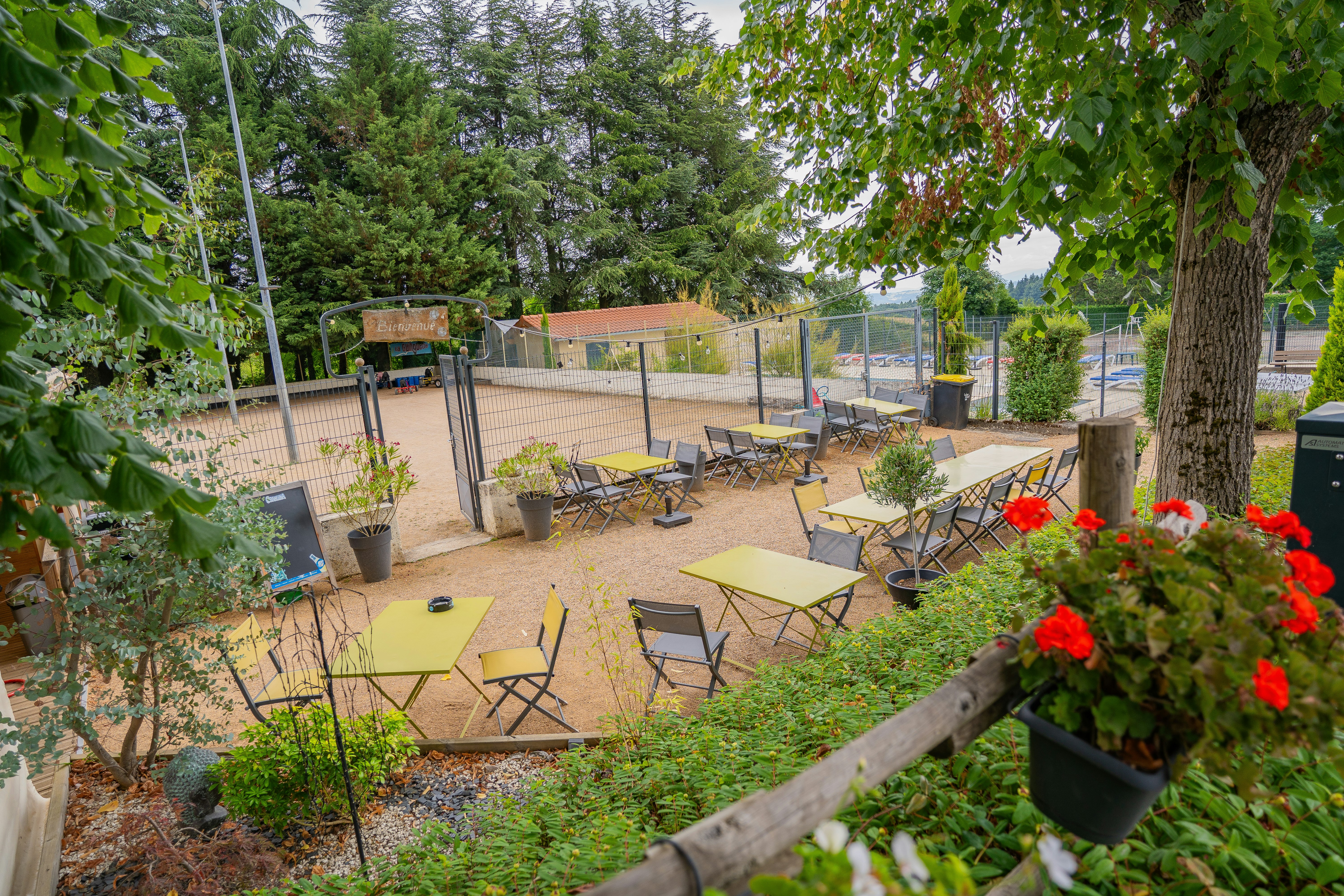 Terrasse mit mehreren Gartenstühlen und Tischen, umgeben von Bäumen und Hecken, mit roten Blumenbeeten im Vordergrund.