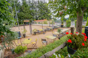 Terrasse mit mehreren Gartenstühlen und Tischen, umgeben von Bäumen und Hecken, mit roten Blumenbeeten im Vordergrund.