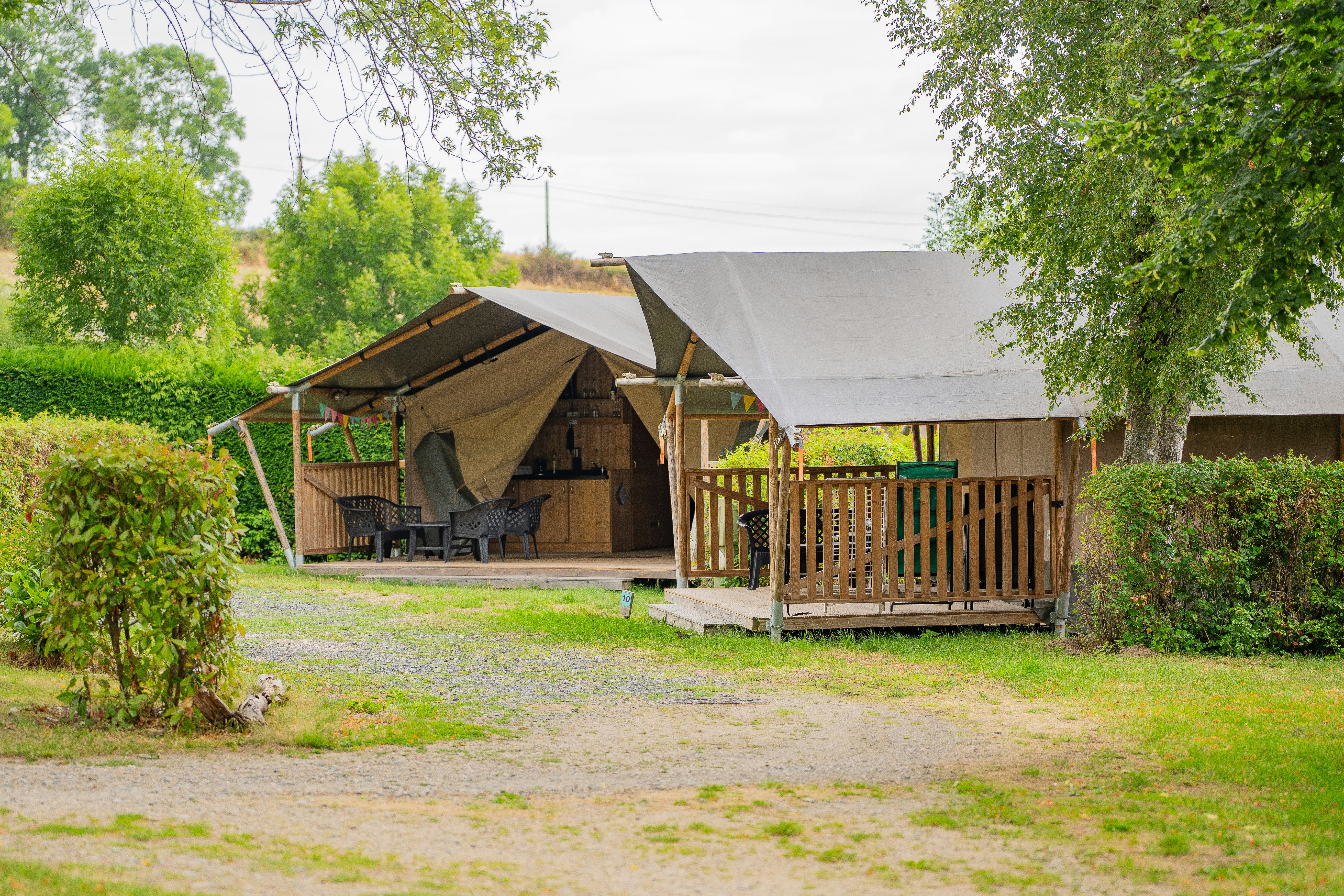 Zelt mit Holzveranda und Sitzbereich auf einem Campingplatz, umgeben von Bäumen und gepflegtem Rasen.