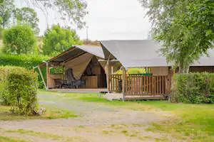 Zelt mit Holzveranda und Sitzbereich auf einem Campingplatz, umgeben von Bäumen und gepflegtem Rasen.