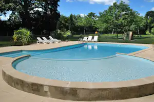 Camping-Pool mit blauem Wasser, Steinterrasse und Liegestühlen im Garten, umgeben von Bäumen und Hecke.