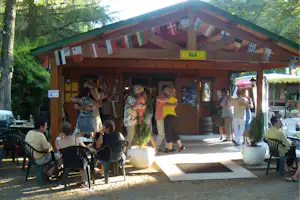 Holzbar am Campingplatz mit bunten Wimpeln, Gäste sitzen an Tischen im Schatten, Bäume im Hintergrund.