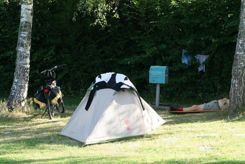 Zelt auf einem grasigen Campingplatz zwischen Bäumen, daneben ein Fahrrad und ein Schild am Wegesrand.