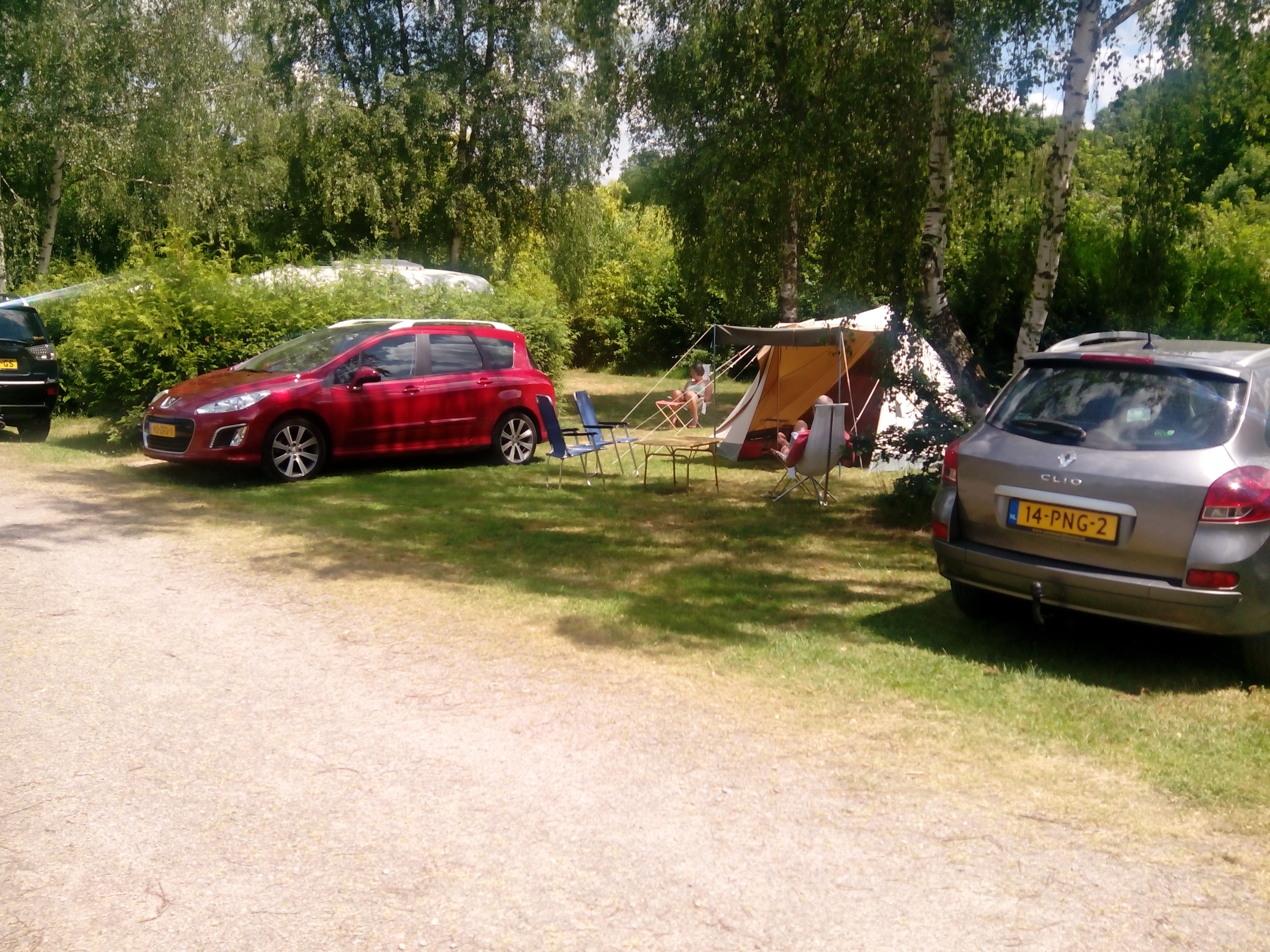 Stellplatz am Campingplatz mit rotem Auto, silbernem Auto und Zelt zwischen Bäumen auf einer Rasenfläche.