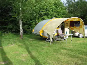 Gelbes Zelt mit Vorzelt und Sitzgruppe auf einem Campingplatz im Grünen, daneben ein Auto und Rasenfläche.