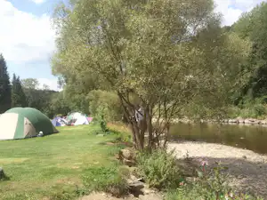 Grünes Campingzelt auf einer Wiese am Flussufer, daneben weitere Zelte und Bäume entlang des Ufers.