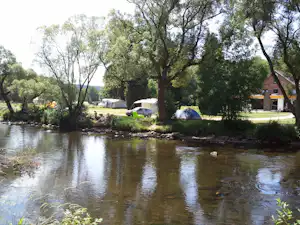 Zelt und Wohnwagen am Flussufer, umgeben von Bäumen, mit Blick auf das ruhige Wasser und einen Weg am Ufer.