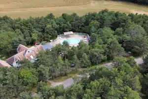 Luftaufnahme vom Campingplatz mit Pool im umzäunten Bereich, Liegen und Sonnenschirm sowie Gebäuden und Wegen im Grünen.