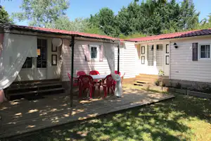 Mobilheim mit Terrasse, Tisch und Stühlen unter einem Sonnendach, umgeben von Rasen und Bäumen im Campingbereich.
