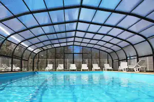 Überdachtes Schwimmbecken mit blauem Wasser, Liegestühlen und Stühlen im Poolbereich, dahinter Zaun und Bäume.