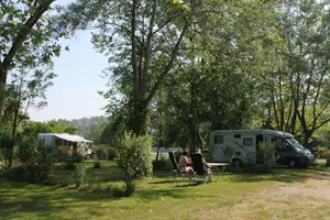 Wohnmobil und Zelt auf einem grünen Campingplatz, daneben Stühle und Tisch im Schatten der Bäume.