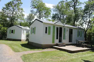 Mobilheim mit Terrasse und Sitzbank, weißes Gebäude mit grünen Akzenten, Rasen und Kiesweg im Campingbereich.