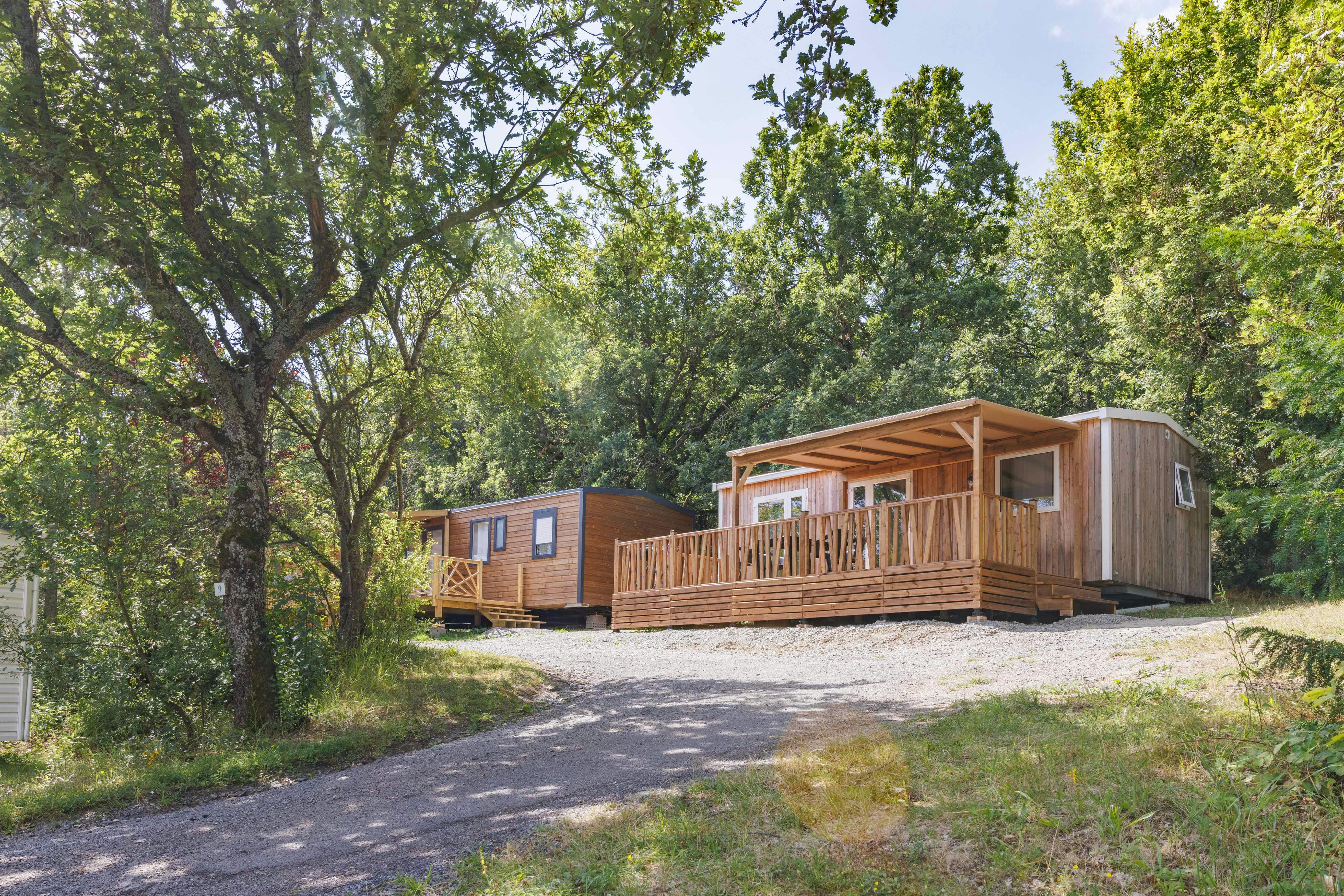Holzhütten und ein Wohnwagen auf einem Campingplatz, mit Holzterrasse und Weg, umgeben von Bäumen im Grünen.