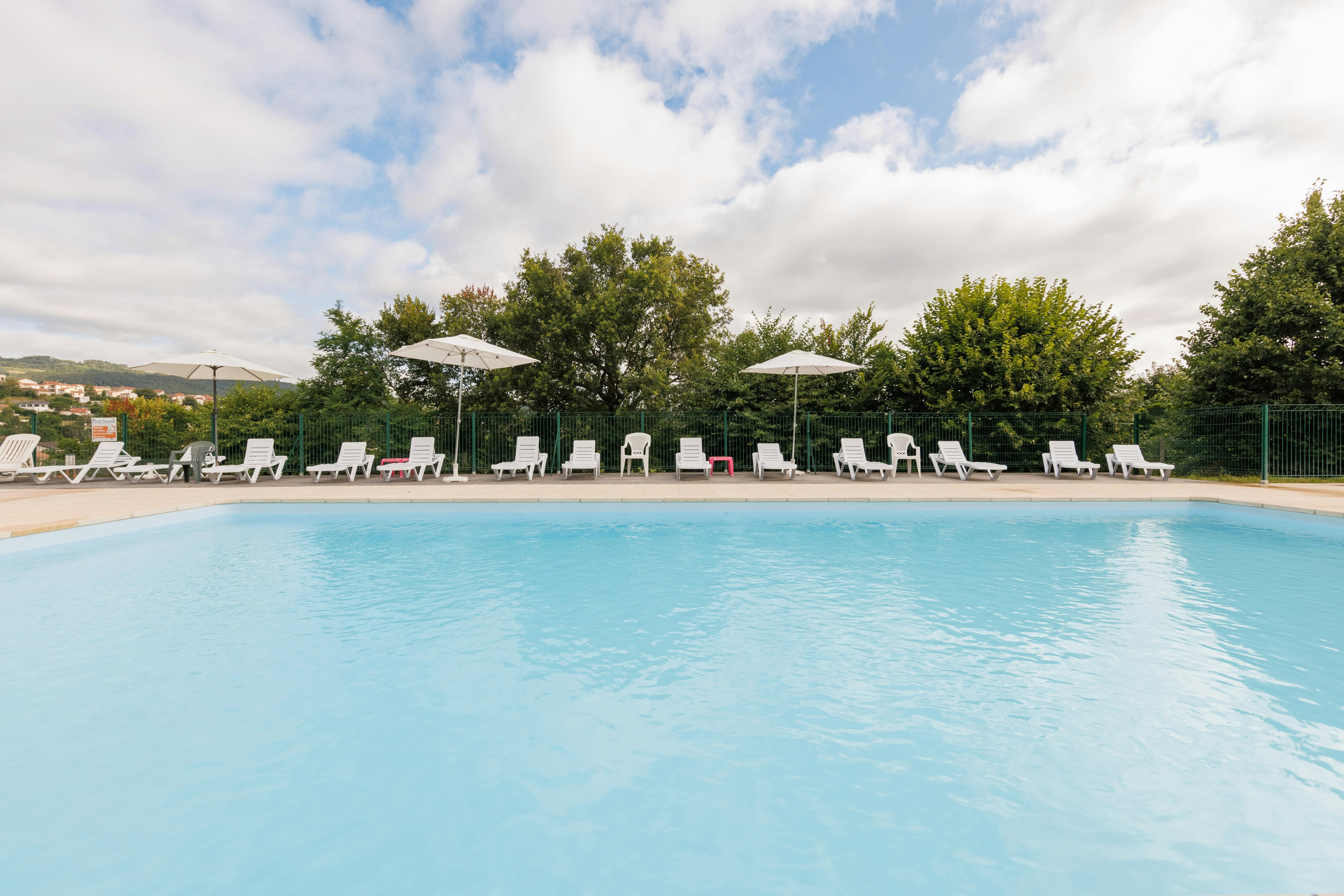 Schwimmbecken mit klarem Wasser, Liegestühlen und Sonnenschirmen am Campingplatz, dahinter Bäume und ein Zaun.