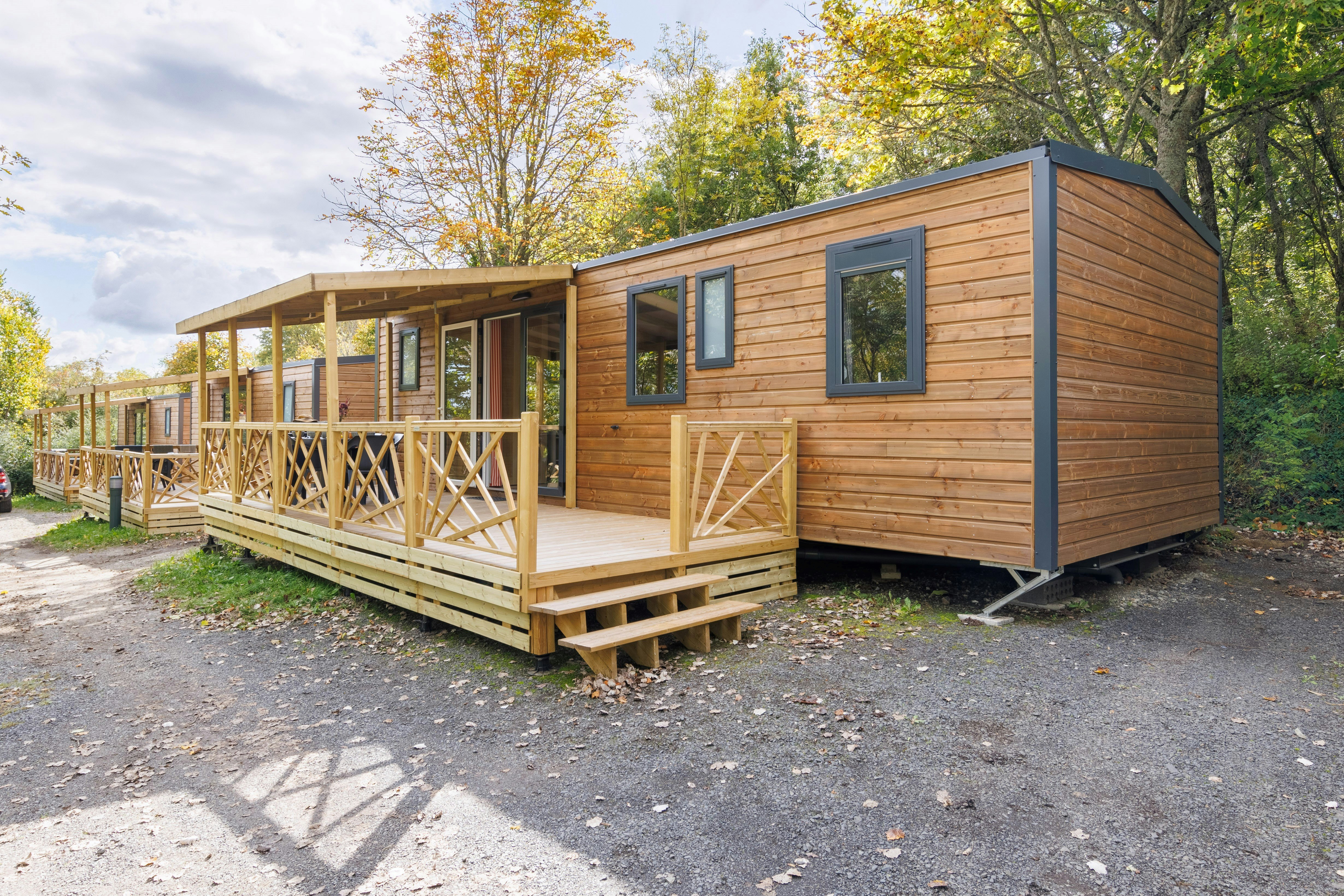 Holz-Blockhaus auf einem Campingplatz mit Veranda und Geländer, umgeben von Bäumen und Kiesweg.