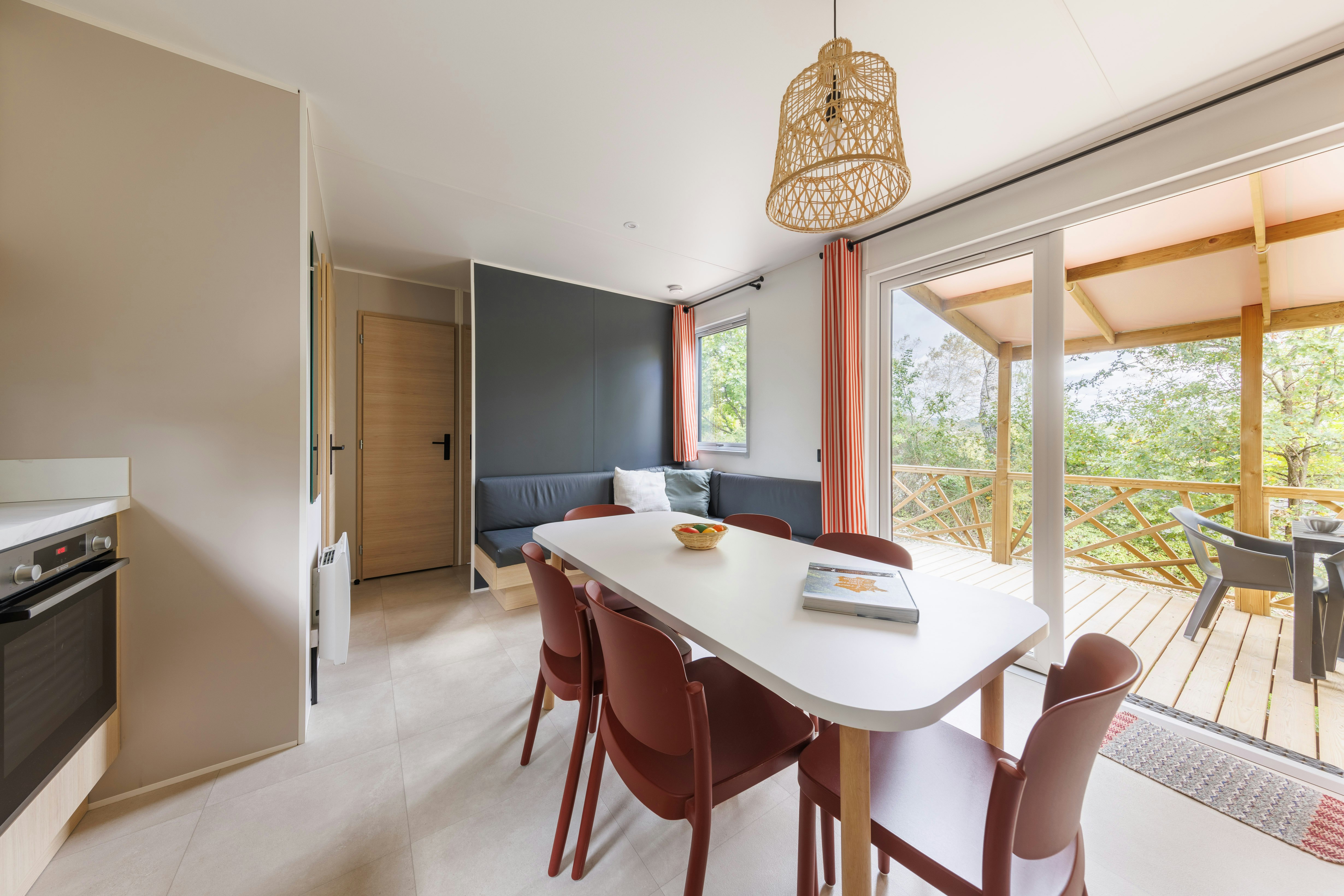 Essbereich in einem Ferienhaus: weißer Tisch mit Stühlen, große Fenster und Holzgeländer auf der Terrasse, Blick ins Grün.