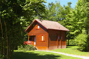 Holzhaus mit rotem Ziegeldach, Fensterläden und kleiner Terrasse im Grünen, umgeben von Bäumen und Rasen.