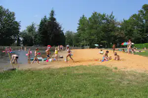 Sandstrand am Fluss mit Familien beim Spielen, bunten Spielzeugen und Sonnenschirmen, im Hintergrund Bäume und Uferweg.