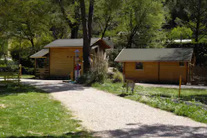 Zwei Holz-Hütten auf einem Kiesweg im Grünen, umgeben von Bäumen und Sträuchern auf dem Campingplatz.