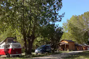Wohnwagen und rote Camping-Van stehen neben einem Baum, dahinter Holzhütten und geparkte Autos auf dem Campingplatz.