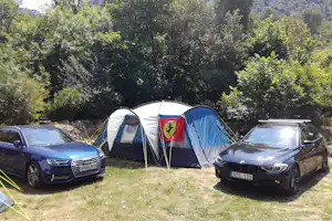 Campingzelt mit blau-weißen Wänden und roter Flagge, davor geparkte Autos auf einer Wiese, umgeben von Bäumen.