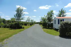 Schotterweg im Campinggelände zwischen Hecken und Bäumen, rechts ein weißes Mobilheim mit blauen Fenstern.
