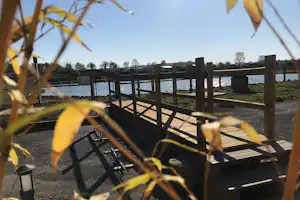 Holzterrasse mit Sitzgelegenheiten und Zaun, im Vordergrund Gräser, dahinter Wasser und Bäume am Campingplatz.