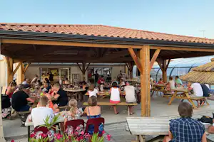 Überdachte Camping-Terrasse mit langen Tischen, vielen Gästen und Holzbänken, daneben ein Gebäude mit Fensterfront.