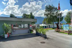 Campingplatz mit Wohnwagen und Wohnmobilen, gepflegtem Rasen, Bäumen und Bergen im Hintergrund unter wolkigem Himmel.