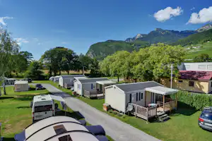 Reihe von Mobilheimen und Wohnwagen auf einem Campingplatz, mit Zufahrtsstraße, Rasen und Bergen im Hintergrund.