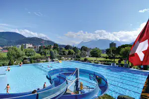 Schwimmbad mit blauer Rutsche, Liegewiese und Bungalows am Hang, im Hintergrund Berge und ein Schweizer Flaggenbanner.