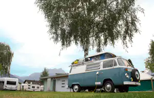 Blaues Wohnmobil mit Dachzelt auf dem Campingplatz, Bäume im Hintergrund und Berge in der Ferne.