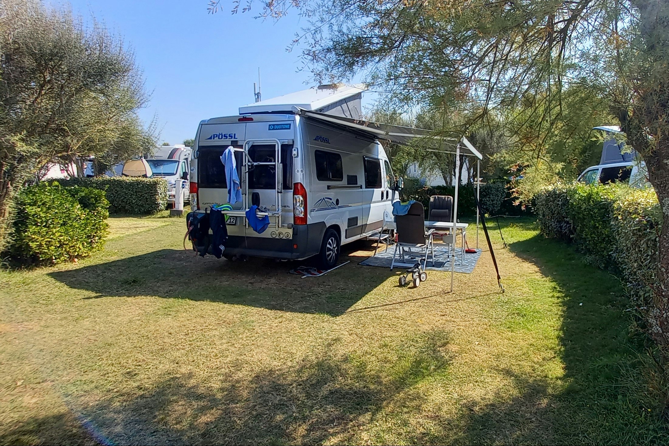 Wohnmobil auf einem Campingplatz-Stellplatz mit Vorzelt und Tisch, umgeben von Rasen, Hecken und Bäumen.