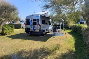 Wohnmobil auf einem Campingplatz-Stellplatz mit Vorzelt und Tisch, umgeben von Rasen, Hecken und Bäumen.