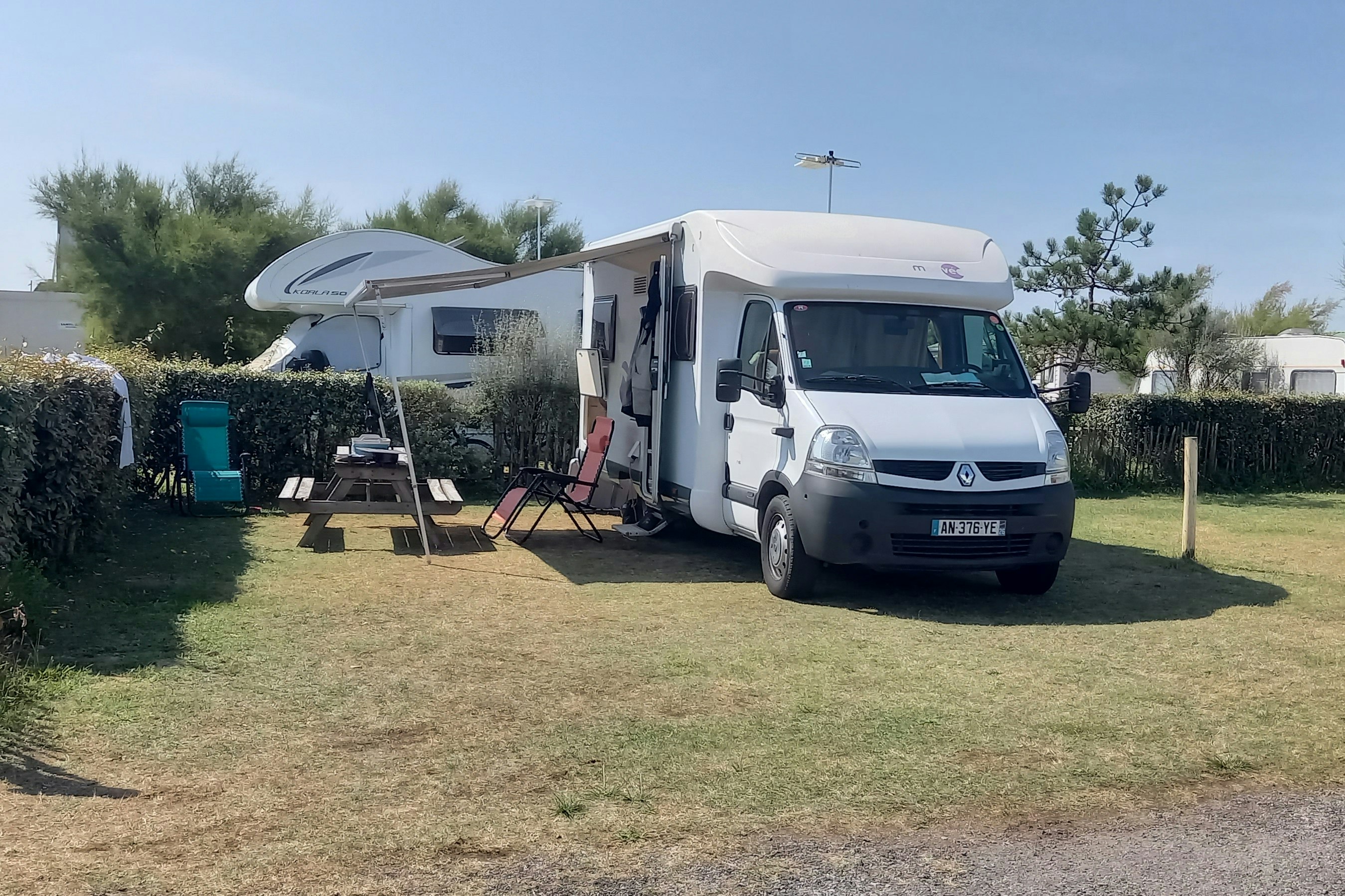 Wohnmobil auf einem Campingplatz mit Rasenfläche, Hecke und Sitzgruppe am Stellplatz, im Hintergrund Bäume und Gebäude.