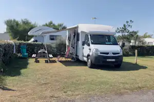 Wohnmobil auf einem Campingplatz mit Rasenfläche, Hecke und Sitzgruppe am Stellplatz, im Hintergrund Bäume und Gebäude.