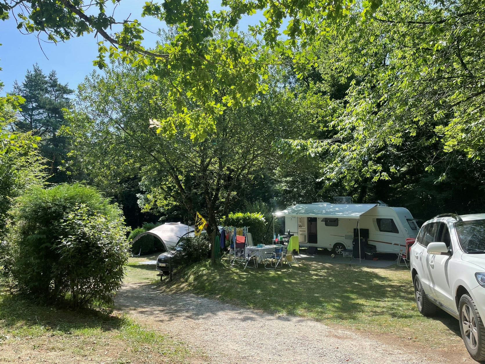 Wohnwagen und Pkw auf einem Campingplatz im Grünen, mit Tisch und Stühlen am Stellplatz zwischen Bäumen.