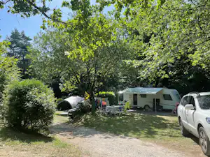 Wohnwagen und Pkw auf einem Campingplatz im Grünen, mit Tisch und Stühlen am Stellplatz zwischen Bäumen.