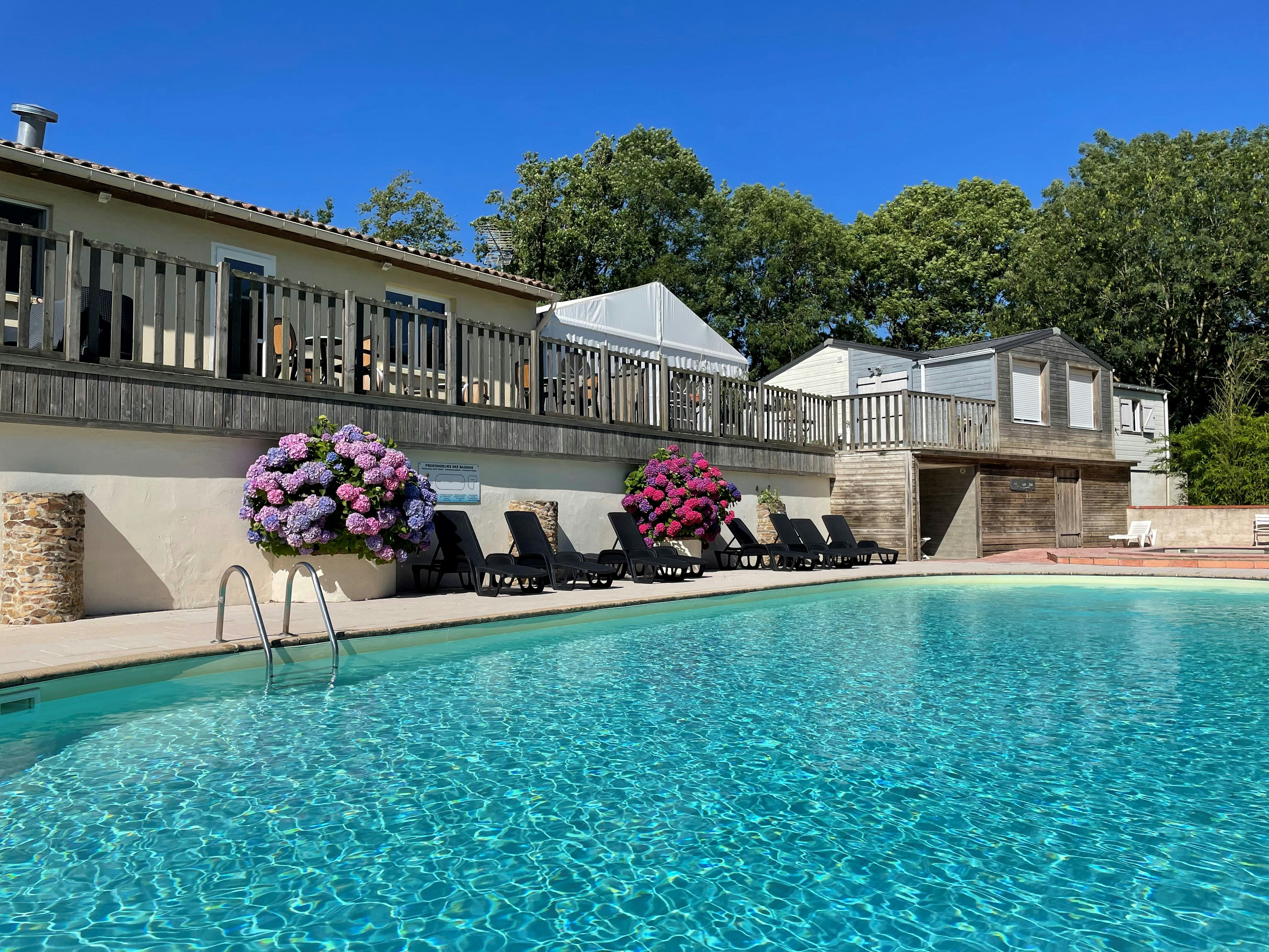 Blauer Campingpool mit Liegen und Sonnenschirmen vor einer Terrasse mit Geländer, dahinter ein Gebäude und Bäume.