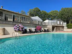 Blauer Campingpool mit Liegen und Sonnenschirmen vor einer Terrasse mit Geländer, dahinter ein Gebäude und Bäume.