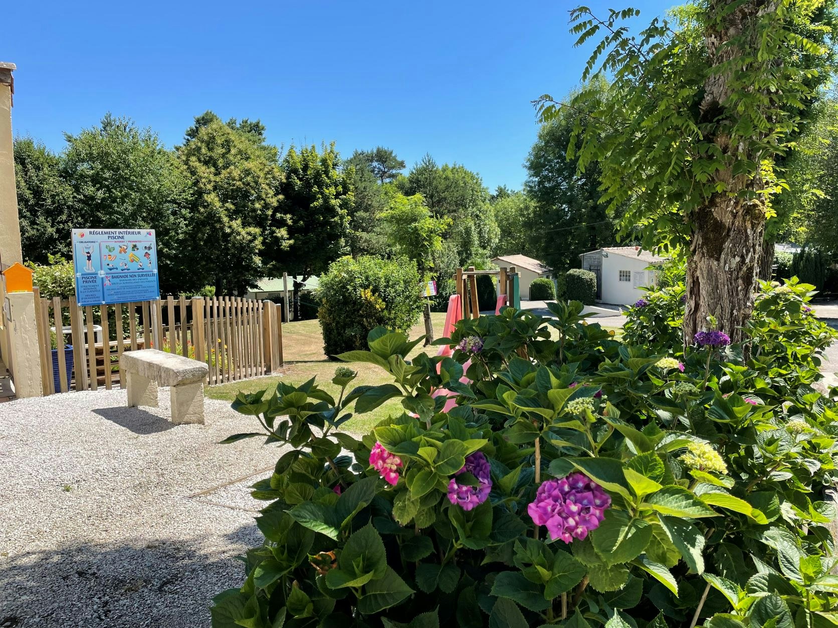 Blumenbeet am Campingweg mit Kiesfläche, Holzzaun und Bäumen, im Hintergrund Wohnwagen und ein Infoposter.