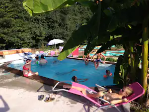 Camping-Pool mit mehreren Liegen und Badegästen, im Vordergrund grüne Blätter und im Hintergrund ein Sonnenschirm.
